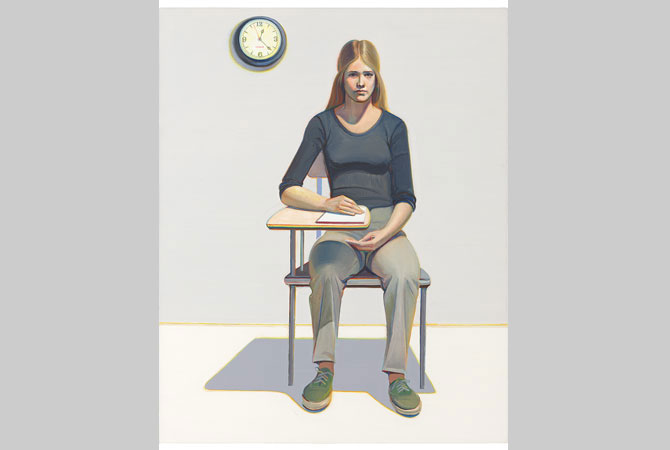 A woman sits in a student’s desk with a pad of paper.