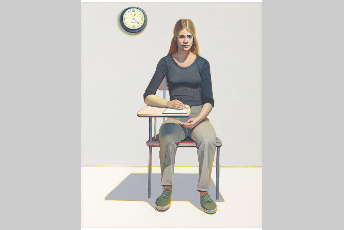 A woman sits in a student’s desk with a pad of paper.