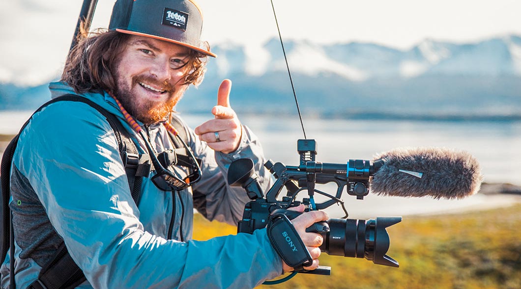Photo of a camera man with a lot of equipment giving thumbs up sign in nature
